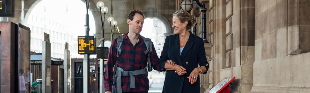 man with whote waliing aid, visually impaired, walking through station with a woman