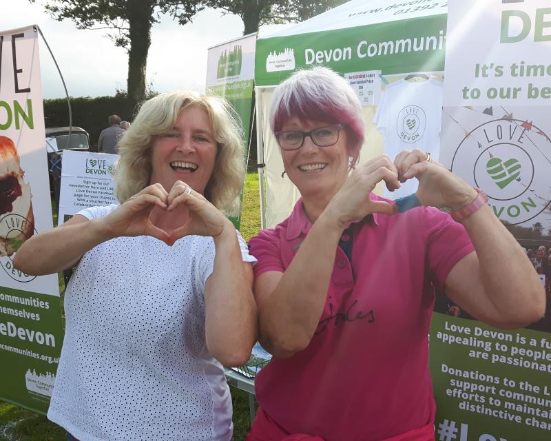 Two people making a heart shape each with both hands in front of Love Devon banners