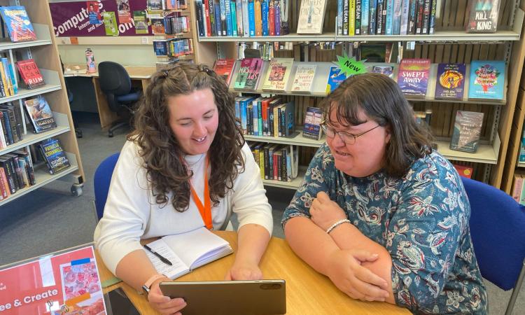 Two people sat in a library setting with an ipad, showing support and advice