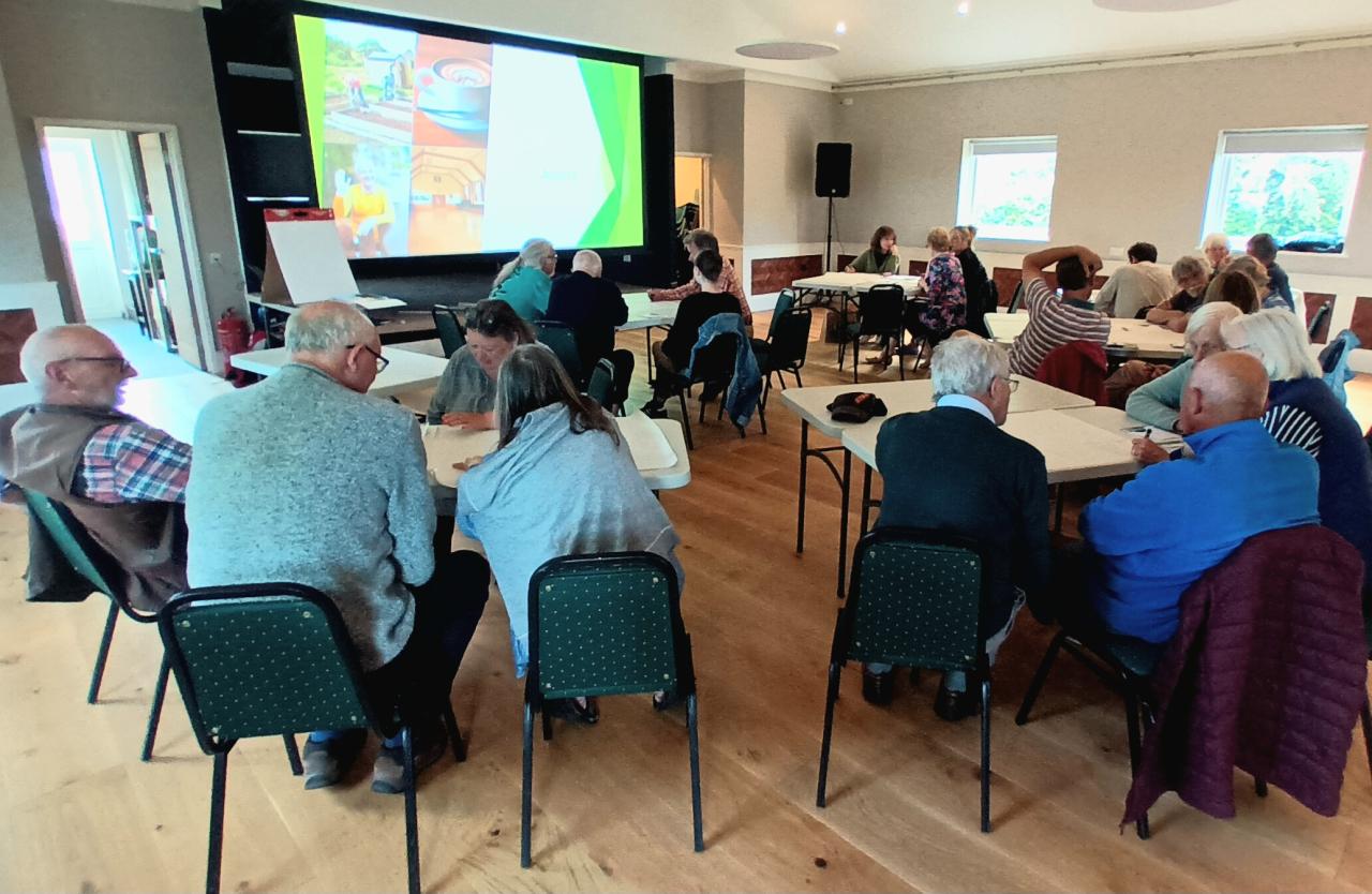 Group of people sat around tables in a village hall, looking at a screen projector