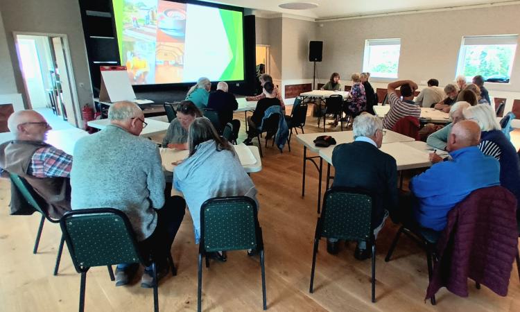 Group of people sat around tables in a village hall, looking at a screen projector